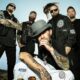 Five men with tattoos posing together outdoors against a blue sky; front man wears a gray hat and black shirt, others in black clothing with motorcycles–style look.