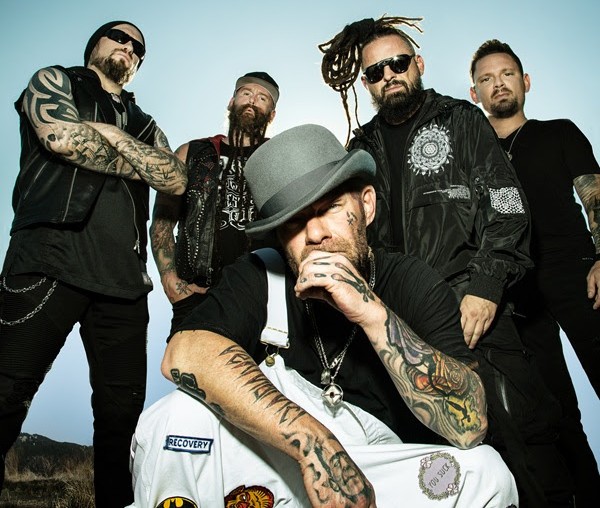 Five men with tattoos posing together outdoors against a blue sky; front man wears a gray hat and black shirt, others in black clothing with motorcycles–style look.
