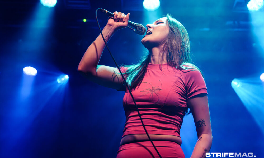 Female vocalist on stage singing into a microphone under blue stage lights, wearing a pink top.