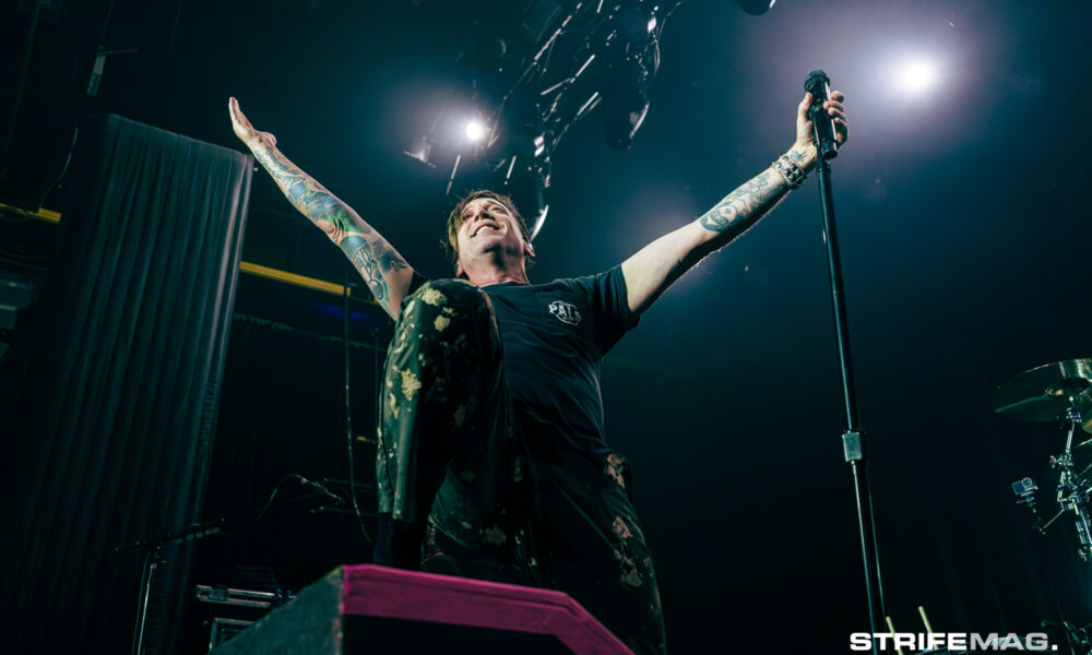 Tattooed lead singer on stage with arms raised, gripping a microphone under bright spotlights.