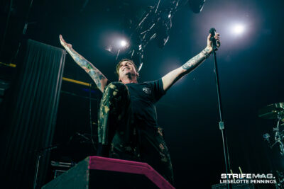 Tattooed lead singer on stage with arms raised, gripping a microphone under bright spotlights.