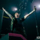 Tattooed lead singer on stage with arms raised, gripping a microphone under bright spotlights.