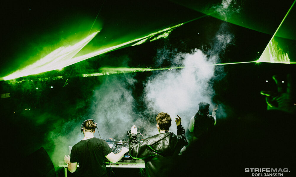 Two DJs stand at a mixing console on a smoky, dark stage as green laser beams sweep overhead in a club.