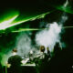 Two DJs stand at a mixing console on a smoky, dark stage as green laser beams sweep overhead in a club.