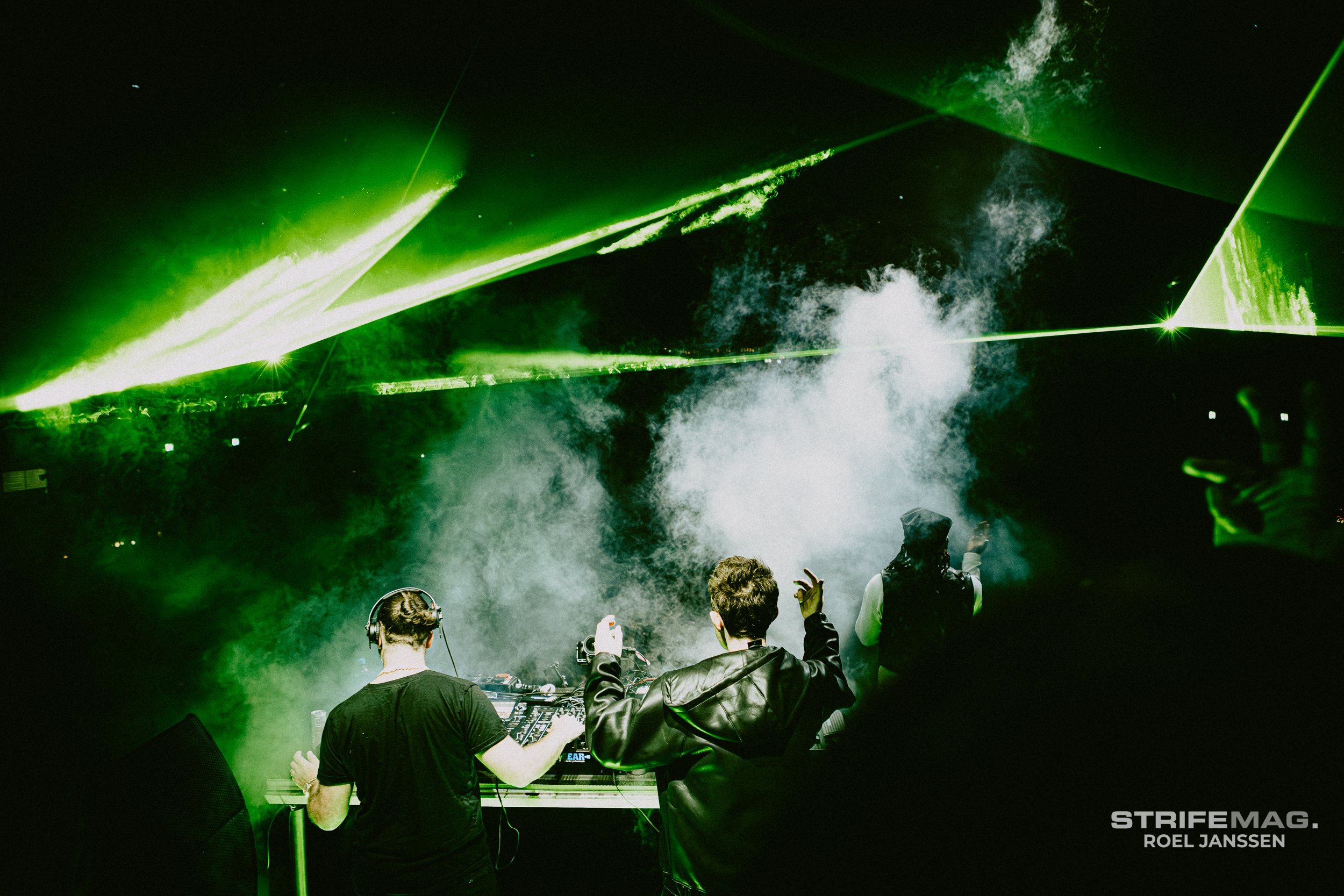 Two DJs stand at a mixing console on a smoky, dark stage as green laser beams sweep overhead in a club.