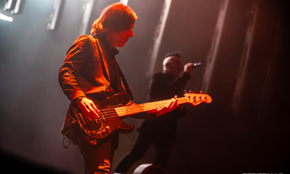 Guitarist playing an electric guitar on stage under warm red lights, with a blurred vocalist in the background.