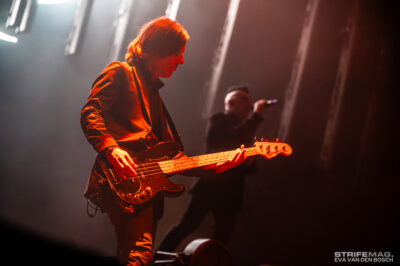 Guitarist playing an electric guitar on stage under warm red lights, with a blurred vocalist in the background.