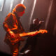 Guitarist playing an electric guitar on stage under warm red lights, with a blurred vocalist in the background.