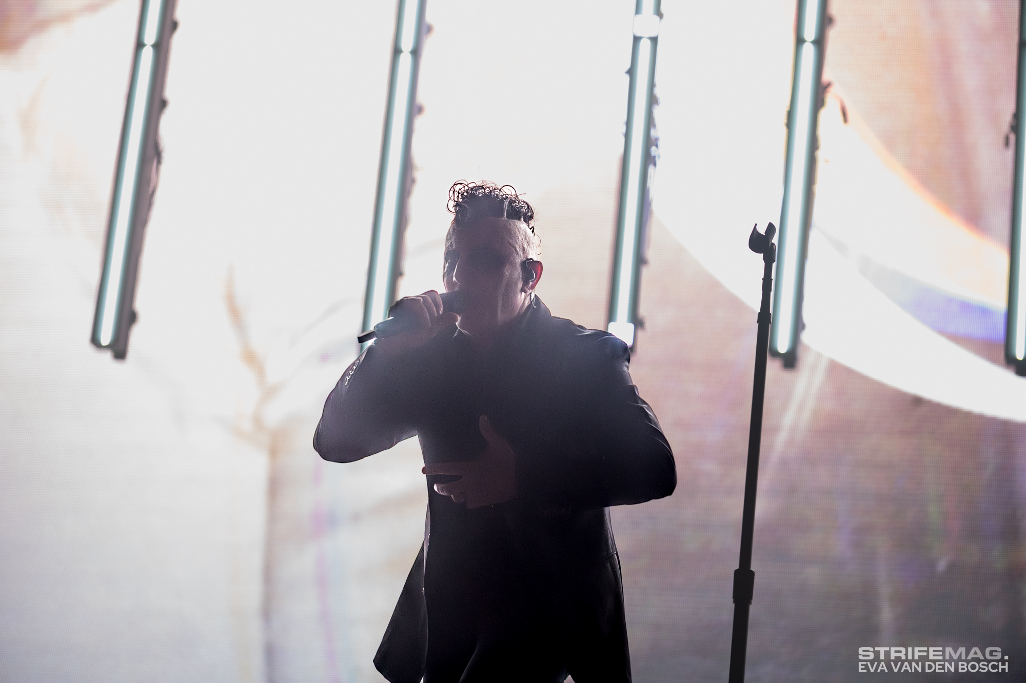 Singer performing on stage, holding a microphone with bright vertical lights behind for a dramatic concert scene.