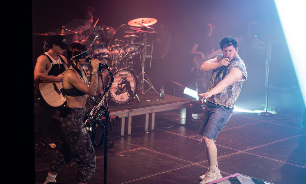 Singer wearing camouflage pants and white tank top sings into a microphone on a smoky stage, with a drummer and guitarist in the background under colored lights.