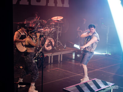 Singer wearing camouflage pants and white tank top sings into a microphone on a smoky stage, with a drummer and guitarist in the background under colored lights.