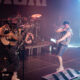 Singer wearing camouflage pants and white tank top sings into a microphone on a smoky stage, with a drummer and guitarist in the background under colored lights.