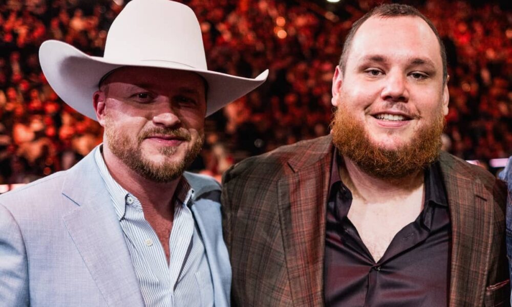 Two men posing at an event; one in a light suit with a white cowboy hat, the other in a plaid blazer.