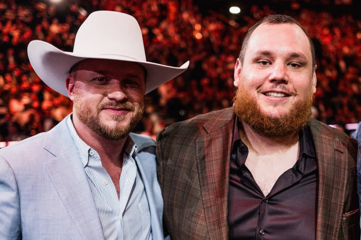 Two men posing at an event; one in a light suit with a white cowboy hat, the other in a plaid blazer.