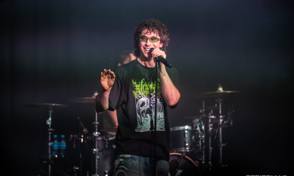 Male rock singer wearing green-tinted sunglasses and a black skull-graphic T-shirt, singing into a handheld microphone on stage.