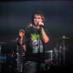 Male rock singer wearing green-tinted sunglasses and a black skull-graphic T-shirt, singing into a handheld microphone on stage.