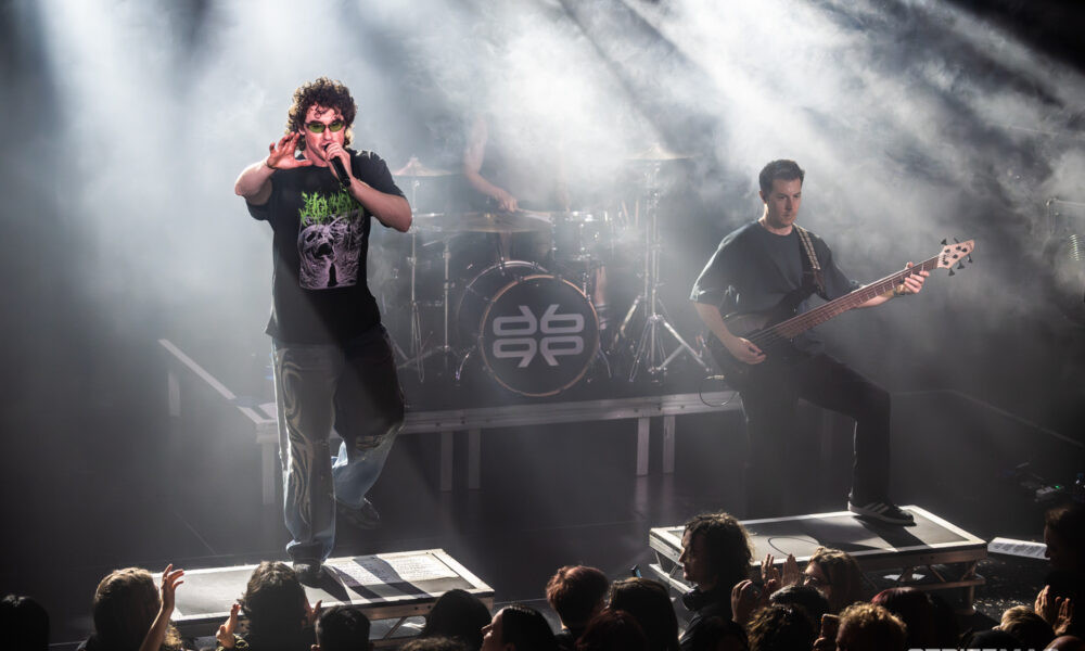 Lead singer with microphone performing on a smoky stage while crowd cheers in front of the platform.