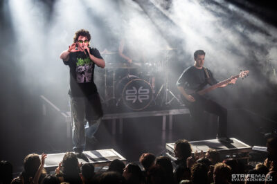 Lead singer with microphone performing on a smoky stage while crowd cheers in front of the platform.