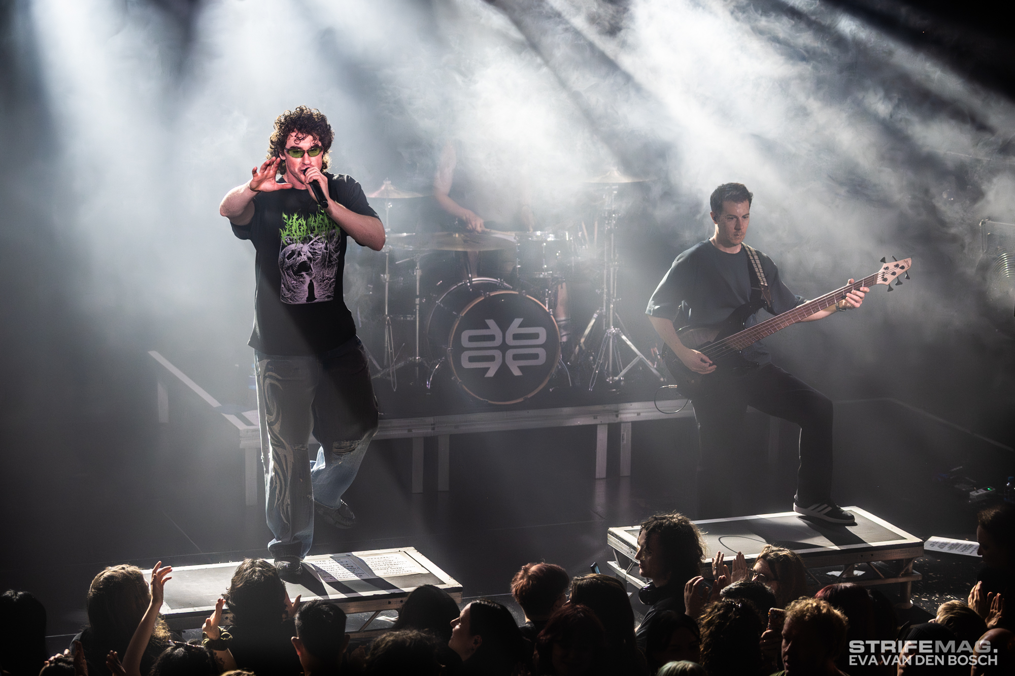 Lead singer with microphone performing on a smoky stage while crowd cheers in front of the platform.