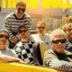 Group of men in sunglasses riding in a bright yellow convertible on a sunny street, posing for a casual band photo.