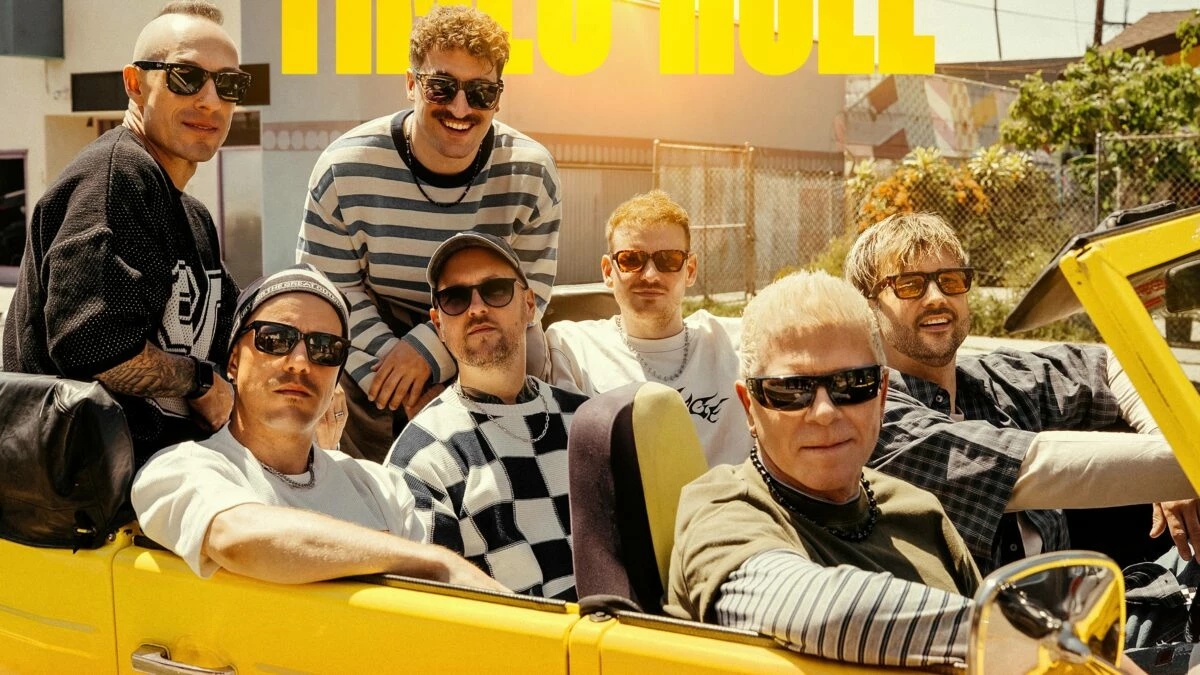 Group of men in sunglasses riding in a bright yellow convertible on a sunny street, posing for a casual band photo.