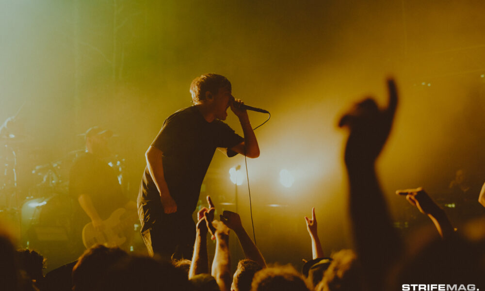 Male singer on stage gripping a microphone as fans reach up, under warm yellow stage lights.