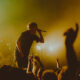 Male singer on stage gripping a microphone as fans reach up, under warm yellow stage lights.