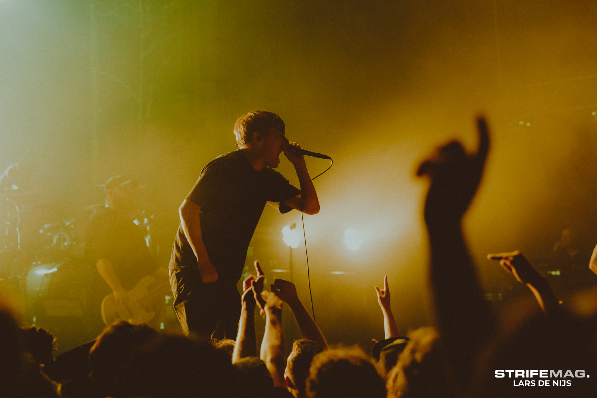 Male singer on stage gripping a microphone as fans reach up, under warm yellow stage lights.