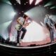 Three musicians perform on a large concert stage, foreground guitarists playing and singing under bright, colorful spotlights with fog and equipment visible on stage.