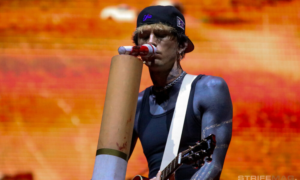 Guitarist in a black cap and sleeveless shirt performs on stage, singing into a cardboard megaphone with an orange blurred crowd behind him.