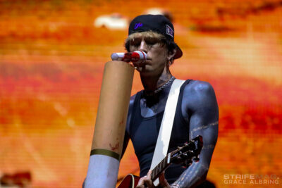 Guitarist in a black cap and sleeveless shirt performs on stage, singing into a cardboard megaphone with an orange blurred crowd behind him.