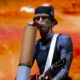 Guitarist in a black cap and sleeveless shirt performs on stage, singing into a cardboard megaphone with an orange blurred crowd behind him.
