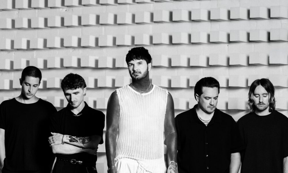 Five men stand in a row against a textured wall; the central figure wears a white mesh top and jewelry, flanked by four casually dressed men in black.