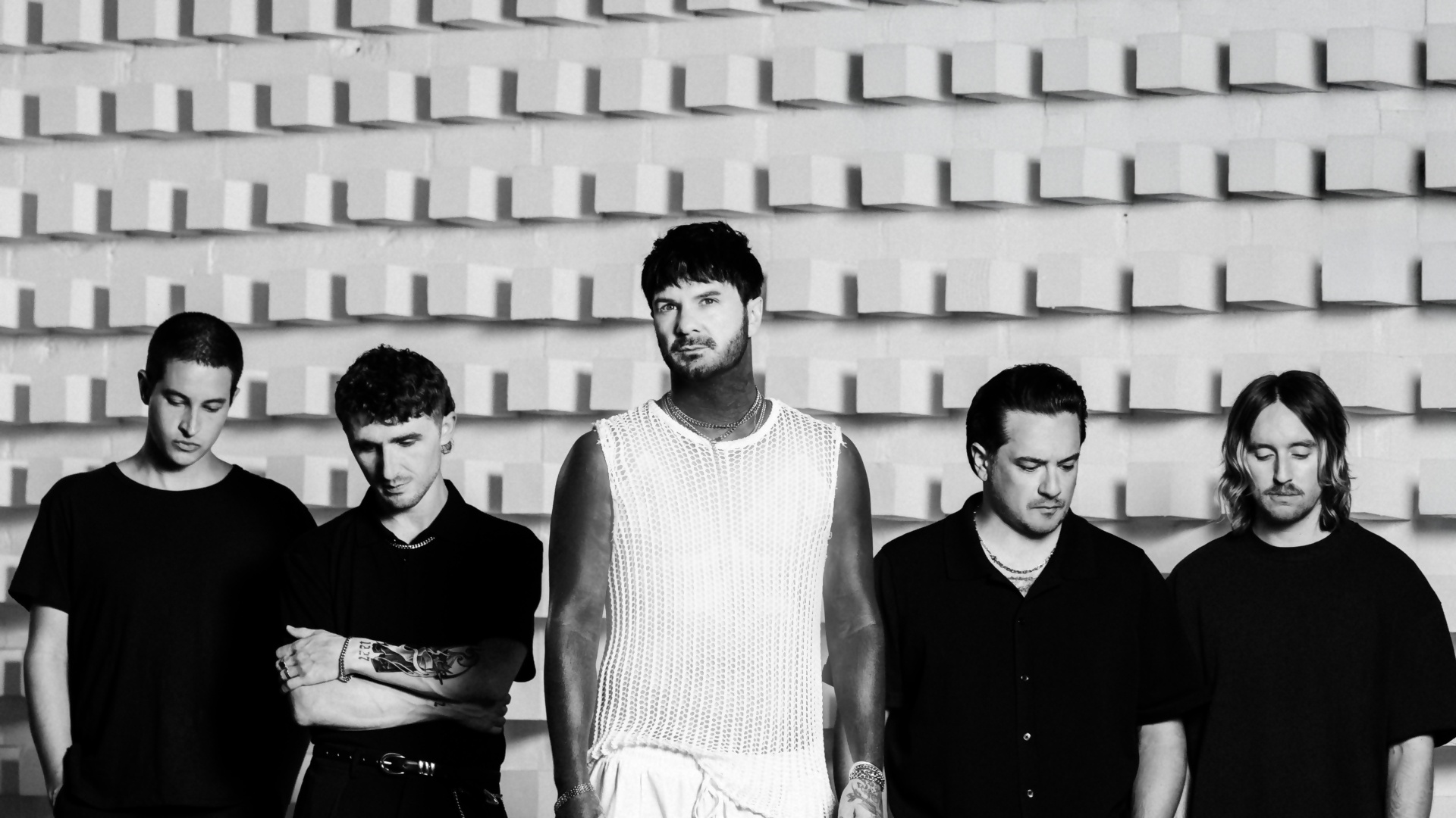 Five men stand in a row against a textured wall; the central figure wears a white mesh top and jewelry, flanked by four casually dressed men in black.