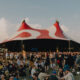 Large crowd at an outdoor festival in front of a red-and-white striped circus tent stage.