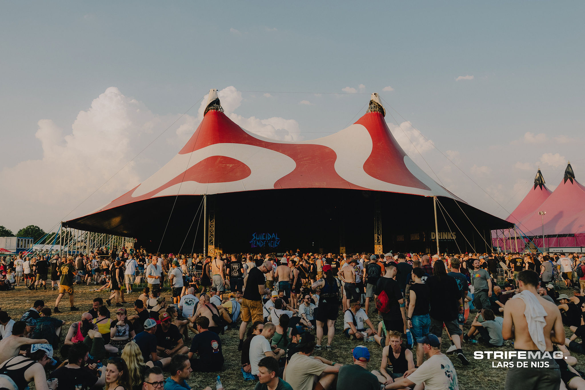 Large crowd at an outdoor festival in front of a red-and-white striped circus tent stage.