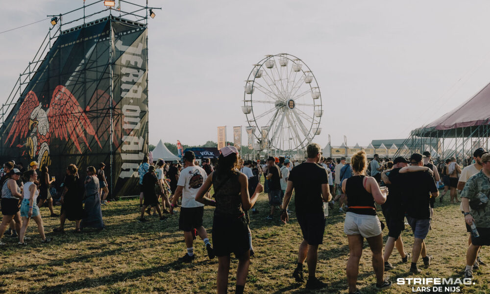 Crowd at an outdoor festival with a large stage on the left and a Ferris wheel in the background.