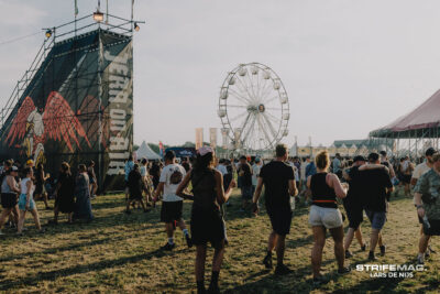 Crowd at an outdoor festival with a large stage on the left and a Ferris wheel in the background.