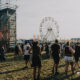 Crowd at an outdoor festival with a large stage on the left and a Ferris wheel in the background.