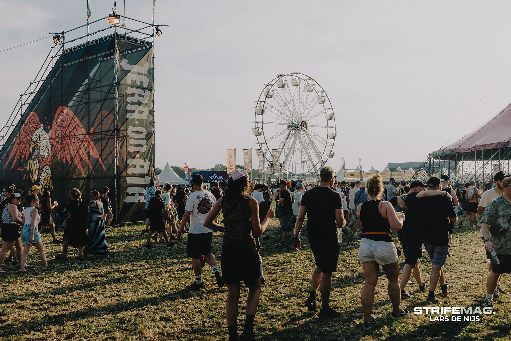Crowd at an outdoor festival with a large stage on the left and a Ferris wheel in the background.