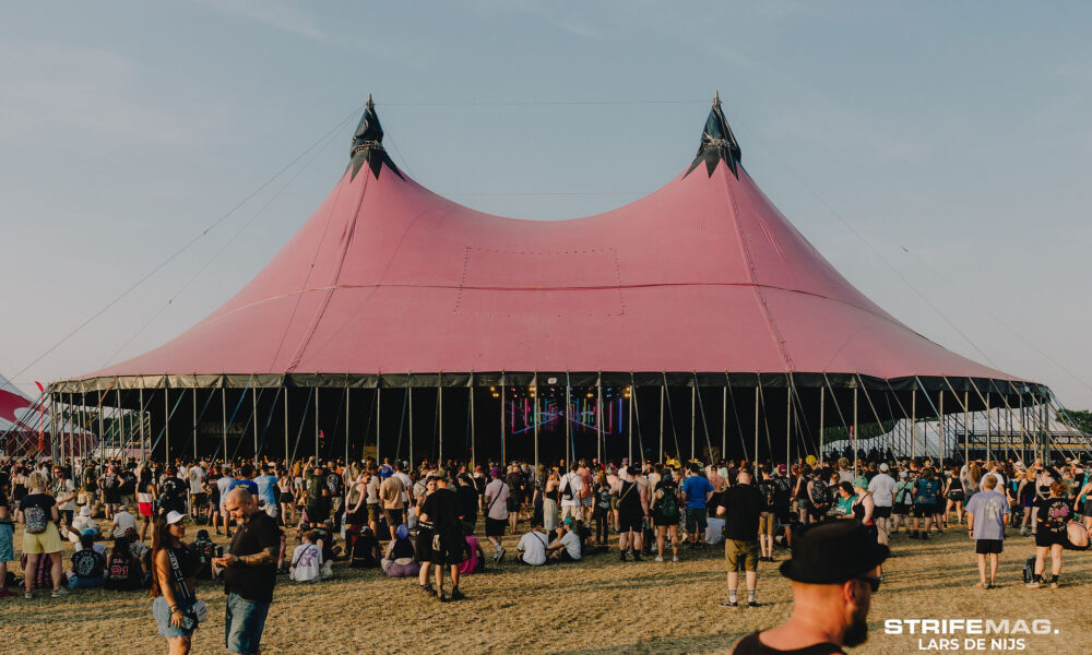 Festival scene with a large pink tent and pointed tops, surrounded by a crowd on a grassy field under a clear sky.