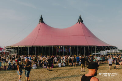 Festival scene with a large pink tent and pointed tops, surrounded by a crowd on a grassy field under a clear sky.