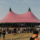 Festival scene with a large pink tent and pointed tops, surrounded by a crowd on a grassy field under a clear sky.
