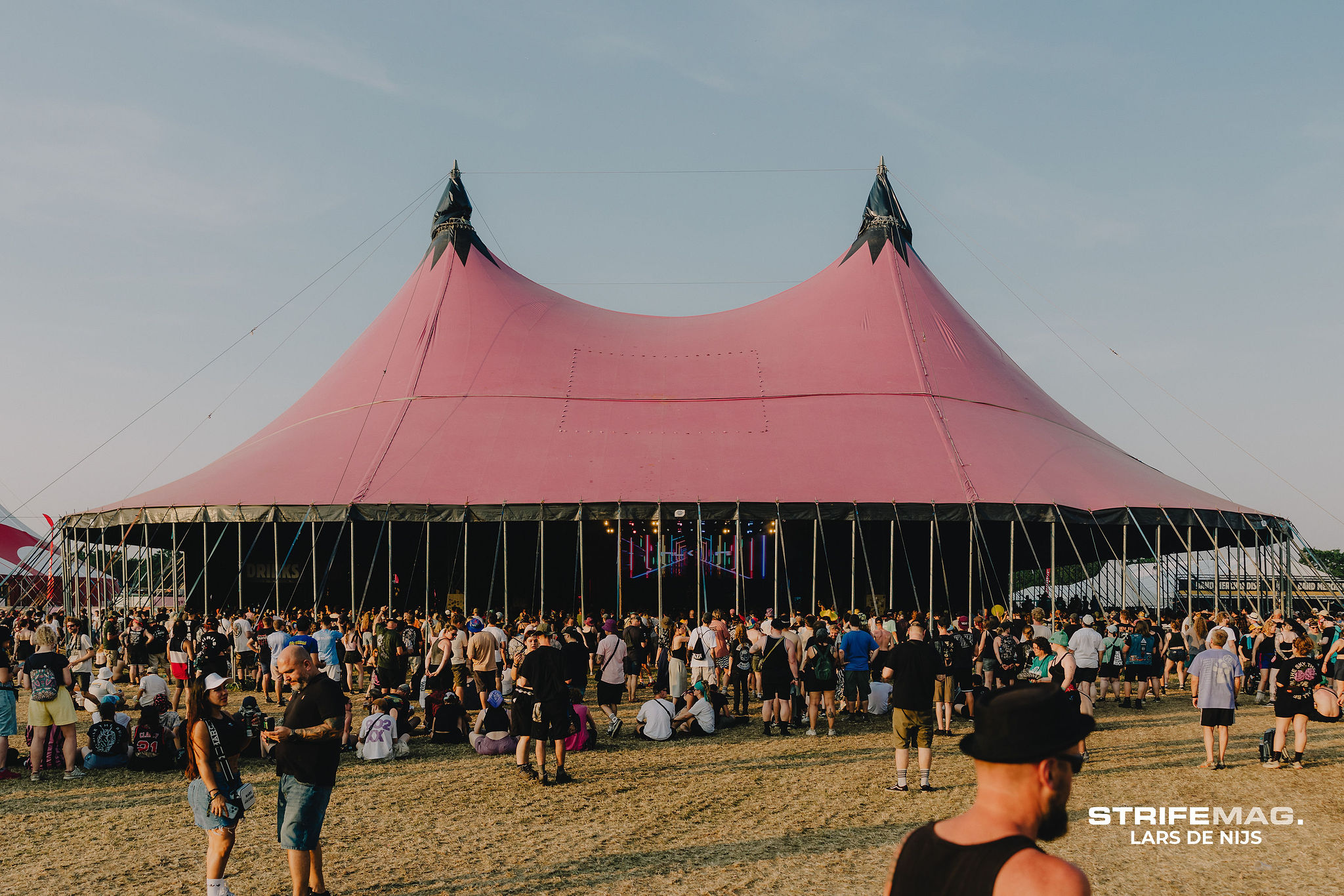 Festival scene with a large pink tent and pointed tops, surrounded by a crowd on a grassy field under a clear sky.