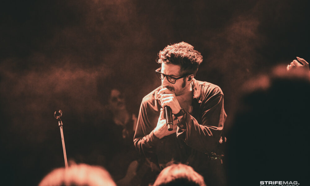 Male singer with glasses performing on a smoky stage, microphone in hand, warm red lighting. Audience silhouettes in foreground.