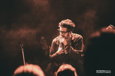 Male singer with glasses performing on a smoky stage, microphone in hand, warm red lighting. Audience silhouettes in foreground.