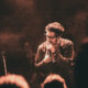 Male singer with glasses performing on a smoky stage, microphone in hand, warm red lighting. Audience silhouettes in foreground.