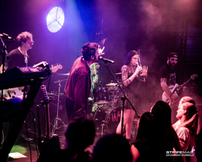 Live concert scene with a male vocalist center stage, a female singer with a microphone at right, guitarist on left, purple stage lighting and visible audience in foreground.
