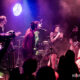 Live concert scene with a male vocalist center stage, a female singer with a microphone at right, guitarist on left, purple stage lighting and visible audience in foreground.