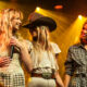 Three female performers on a stage bathed in warm yellow lights, one holding a microphone and smiling at two others.